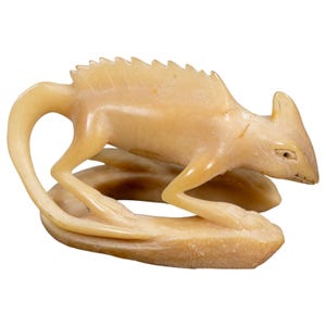 May include: A light beige, carved stone figurine depicting a lizard-like creature with a serrated dorsal ridge, a long curved tail, and a pointed head. The creature is positioned in a crouched stance atop a smooth, oval-shaped base.