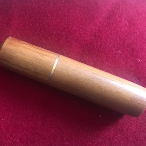 May include: A wooden toothpick holder with a light brown finish and a thin, metallic band. The cylindrical holder has a rounded top and is set against a red background.