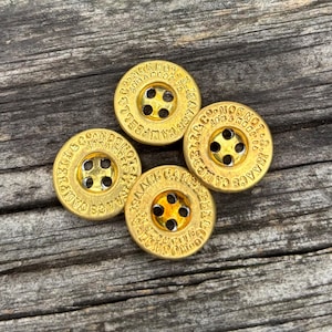 Puede incluir: Un conjunto de tres botones circulares de metal en tono dorado, cada uno con cuatro orificios centrales y texto en relieve alrededor del borde que dice Isaac Campbell & Co. London. Los botones tienen un acabado metálico y un borde elevado.
