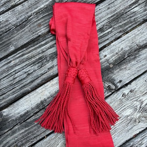 Puede incluir: Una faja roja con largos flecos. La faja está hecha de una tela lisa y tiene un diseño tradicional.