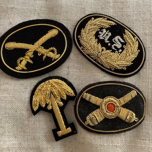 Puede incluir: Cuatro parches bordados con fondos de fieltro negro y diseños de hilo dorado. Un parche presenta un par de espadas cruzadas, otro una palmera, otro una corona con el texto "R.S." y el último un par de correas cruzadas con un centro rojo.