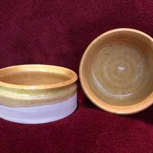 May include: Two handmade ceramic bowls. One is a shallow, open bowl, and the other is a small, lidded container. The bowls are tan with a white base. The bowls are likely used for food or storage.
