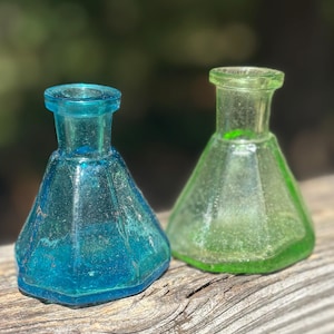 Puede incluir: Dos botellas de vidrio antiguas, una turquesa y otra verde, están una al lado de la otra. Las botellas tienen una base hexagonal y un cuello acampanado. La botella turquesa tiene un aspecto ligeramente desgastado, mientras que la botella verde es más translúcida.