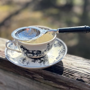 May include: A white teacup with a black and white floral design and a silver tea strainer with a black handle. The teacup is sitting on a matching saucer.