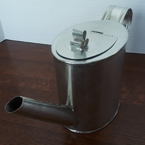 Könnte beinhalten: Eine silberfarbene Metallkanne mit Ausguss, Henkel und Klappdeckel. Der Deckel hat einen dekorativen Verschluss. Die Kanne ist zylindrisch mit ovalem Boden und steht auf einer dunklen Holzoberfläche. Der Ausguss ist abgewinkelt und ragt aus dem Körper heraus.