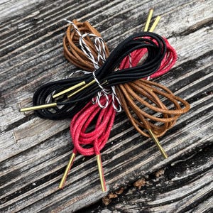 May include: A collection of shoelaces in various colors, including black, red, and brown, with gold-colored aglets. The laces are arranged in a starburst pattern on a weathered wooden surface. The shoelaces are new.