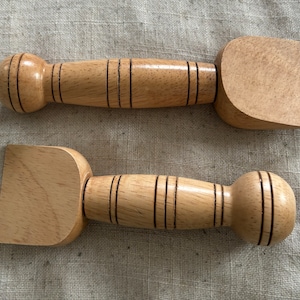 May include: Two wooden tools with rounded handles and flat, slightly curved blades. The tools are made of light brown wood with dark brown lines.