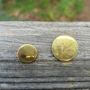 May include: Two round, gold-colored metal buttons on a weathered wooden surface.