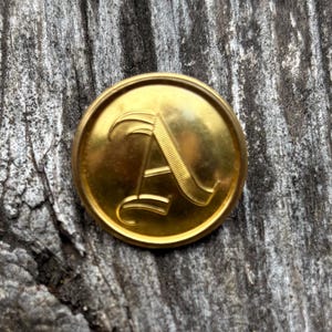 Op de afbeelding: Een goudkleurige knoop met de hoofdletter "A" in een decoratief lettertype. De knoop is rond en heeft een glanzend, reflecterend oppervlak. De knoop ligt op een verweerde, grijze houten achtergrond.