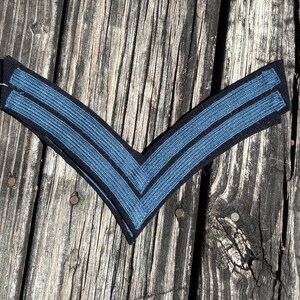 May include: A V-shaped military rank insignia patch featuring two parallel blue chevron stripes embroidered onto a black fabric base. The chevron design is pointed downwards, showcasing a textured, ribbed appearance on the blue stripes.