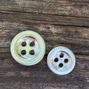 May include: Two iridescent, round buttons with four holes each. The buttons have a pearlescent sheen, reflecting colors of yellow, green, and pink. They are set against a weathered, brown wooden surface, highlighting their unique color.