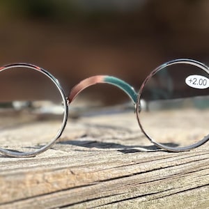 May include: Round, silver-framed reading glasses with a +2.00 magnification. The bridge of the glasses has a brown and green pattern. The glasses are resting on a weathered wooden surface, with a blurred green background.