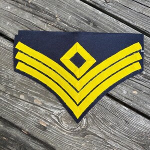 Puede incluir: Un parche de tela azul marino oscuro con tres franjas de cheurón amarillas horizontales dispuestas en forma de V, con un emblema amarillo en forma de diamante centrado sobre la franja superior. El parche tiene un borde inferior puntiagudo.