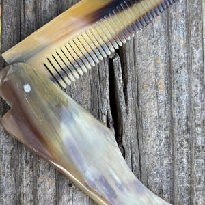 Puede incluir: Un peine plegable hecho de cuerno natural, con una gama de colores que va del amarillo claro al marrón oscuro. Los dientes del peine son negros y el mango tiene un acabado liso y pulido. El peine está abierto, mostrando toda su longitud.