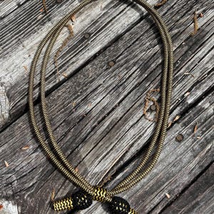 Puede incluir: Un collar de cuerda dorada y negra con un diseño trenzado. El collar está en bucle y tiene detalles en negro y dorado en los extremos. El collar está sobre una superficie de madera desgastada.