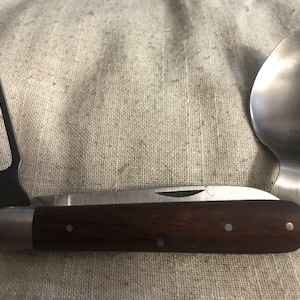 May include: A vintage multi-tool with a spoon, fork, and knife. The spoon and fork are made of stainless steel, and the knife has a wooden handle. The tool is open, displaying all its components.