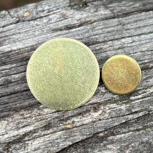 May include: Two brass metal discs of different sizes. The larger disc is approximately 2 inches in diameter, and the smaller disc is about 0.75 inches across. Both have a brushed texture and a golden hue, set against a weathered wood background.