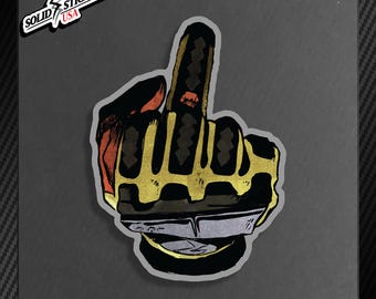 May include: A sticker featuring a stylized hand giving the middle finger. The hand is black and gold, with red accents. The sticker has the text "SOLID STICKERS USA" and "SOLID BLACK 01 ART".