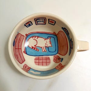 May include: A ceramic mug featuring an interior hand-painted illustration of a bedroom scene. The design includes a white cat sleeping on a blue bed, a red dresser, a rug, and wall art depicting fish and a paw print, all rendered in red and blue.