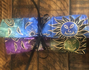 May include: A fabric pouch with a black ribbon tied around it. The pouch is made of a blue and purple fabric with a gold sun design. The sun has a smiling face with eyelashes.