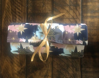 May include: A rolled-up fabric pouch with a gold ribbon tied around it. The pouch is decorated with a pattern of gold stars on a blue, pink, and white background.