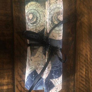 May include: A rolled up fabric pouch with a celestial design featuring constellations and planets. The pouch is tied with a black ribbon.