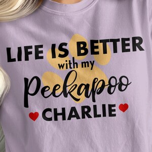Può includere: Una maglietta lilla chiaro con la scritta "LIFE IS BETTER with my Peekapoo CHARLIE" in corsivo nero e una stampa di impronta di zampa. Due cuori rossi affiancano il nome. La maglietta è in stile casual.