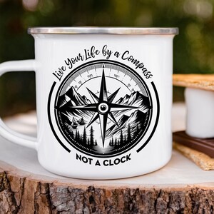 Op de afbeelding: Witte emaille campingmok met een zwart kompasontwerp en de tekst "Live Your Life by a Compass Not a Clock".