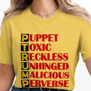 Puede incluir: Camiseta amarilla con la palabra "TRUMP" escrita verticalmente en cuadros negros. Cada letra está coronada con una palabra roja: "Marioneta", "Tóxico", "Imprudente", "Desquiciado", "Malicioso", "Perverso".