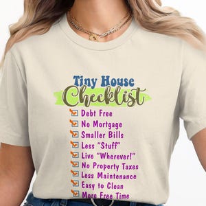 May include: A white t-shirt with a green, brown, and blue graphic that says "Tiny House Checklist" with a list of reasons to live in a tiny house. The list includes: Debt Free, No Mortgage, Smaller Bills, Less "Stuff", Live "Wherever!" No Property Taxes, Less Maintenance, Easy to Clean, More Free Time.