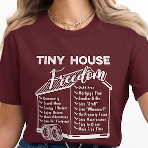 May include: Burgundy t-shirt with the words "TINY HOUSE Freedom" in white. A graphic of a tiny house on wheels is below the text, with a list of benefits. The shirt is made of a soft material.