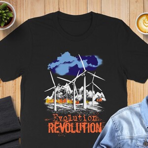 May include: Black t-shirt featuring a graphic of wind turbines, mountains, and a blue cloud. The text "Evolution Revolution" is printed in orange. A cup of coffee and a small plant are in the background.