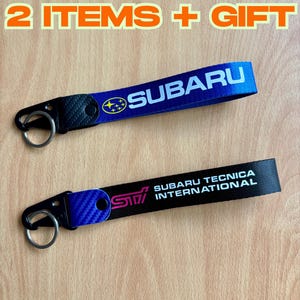 Puede incluir: Dos llaveros Subaru. Uno es azul con el logo de Subaru y el otro es negro con el logo STI y el texto "Subaru Tecnica International". Ambos tienen un mosquetón negro y un llavero.