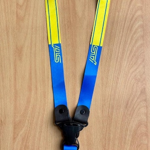 Könnte beinhalten: Ein gelb-blaues Schlüsselband mit den Buchstaben "STI" auf blauem Grund. Das Schlüsselband hat einen schwarzen Verschluss und einen silbernen Schlüsselring. Der Hintergrund ist eine hellbraune Holzoberfläche.