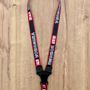 Puede incluir: Cordón negro y rojo con el texto "YOSHIMURA" en blanco. El cordón tiene un cierre negro y un anillo metálico. El material es tejido. Diseñado para llevar alrededor del cuello.