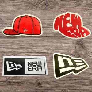 Puede incluir: Cuatro pegatinas con el logotipo de New Era y diseños de gorras de béisbol. Las pegatinas incluyen una gorra de béisbol roja, una gorra roja con el texto "NEW ERA", una pegatina rectangular en blanco y negro con el nombre de la marca y un logotipo verde y blanco.