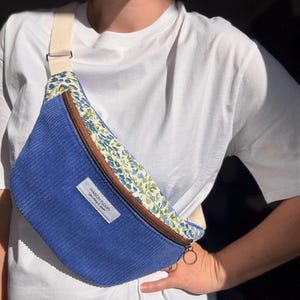 Pode incluir: Cinto de cintura de veludo cotelê azul com um detalhe floral e uma alça creme. Possui um zíper marrom e uma etiqueta com a inscrição "MAISON FLOURY Upcycling & stuff".