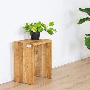 Könnte beinhalten: Ein Beistelltisch aus Naturholz mit quadratischer Oberseite und offenen Seiten. Auf der Oberseite steht ein kleiner schwarzer Topf mit einer grünen Pflanze. Im Hintergrund eine große Topfpflanze mit grünen Blättern.