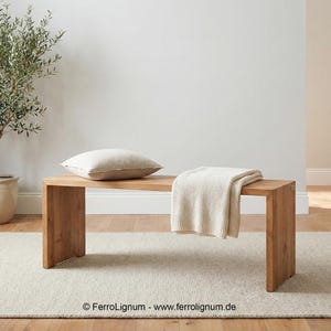 Könnte beinhalten: Eine helle Holzbank mit einem cremefarbenen Kissen und einer Decke. Links steht ein Olivenbaum im Topf. Die Bank steht auf einem hellbeigen Teppich, mit einer hellgrauen Wand im Hintergrund.