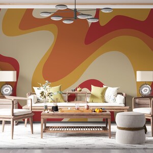 Op de afbeelding: Een woonkamer met een witte bank en twee stoelen, een salontafel en een ronde poef. De muur achter de meubels is geschilderd met een retro-stijl abstract patroon in tinten oranje, geel en rood.