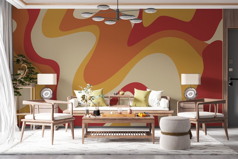 Op de afbeelding: Een woonkamer met een witte bank en twee stoelen, een salontafel en een ronde poef. De muur achter de meubels is geschilderd met een retro-stijl abstract patroon in tinten oranje, geel en rood.