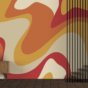 Op de afbeelding: Een kamer met een muur bedekt met een retro-behang met oranje, gele en rode abstracte vormen. Een houten plank met boeken en een potplant staat voor de muur. Een zwarte metalen scheidingswand met verticale latten bevindt zich aan de rechterkant van de afbeelding.