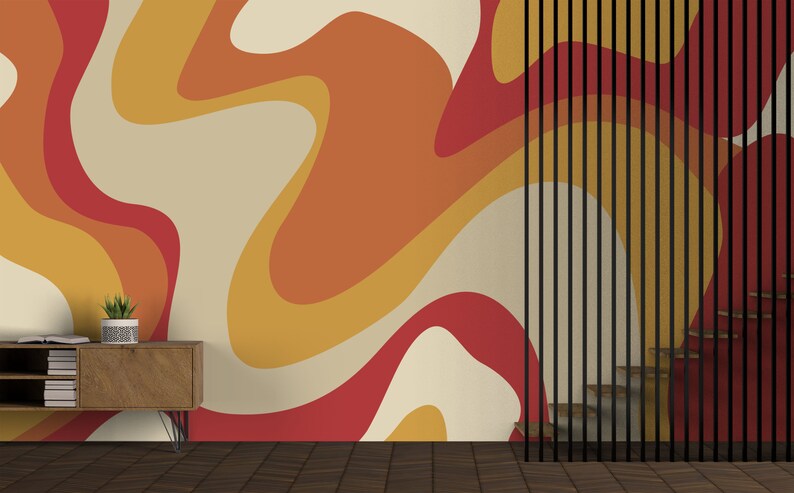 Op de afbeelding: Een kamer met een muur bedekt met een retro-behang met oranje, gele en rode abstracte vormen. Een houten plank met boeken en een potplant staat voor de muur. Een zwarte metalen scheidingswand met verticale latten bevindt zich aan de rechterkant van de afbeelding.