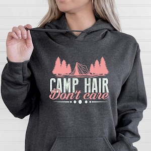 K&ouml;nnte beinhalten: Dunkelgrauer Kapuzenpullover mit Fronttasche und Kordelzug. Die Grafik auf der Vorderseite zeigt ein rosa Zelt und B&auml;ume mit dem Text "CAMP HAIR Don't care."