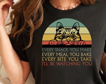 Puede incluir: Camiseta negra con un diseño de gato sobre un fondo de puesta de sol. El texto dice: "EVERY SNACK YOU MAKE, EVERY MEAL YOU BAKE, EVERY BITE YOU TAKE, I'LL BE WATCHING YOU."