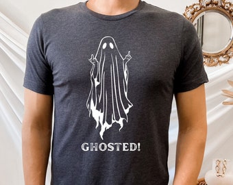 May include: Dark gray t-shirt featuring a white graphic of a ghost giving the middle finger. The word "GHOSTED!" is printed below the ghost. The shirt has short sleeves and a crew neck.