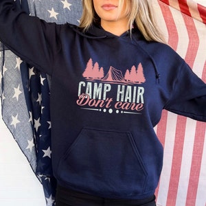 K&ouml;nnte beinhalten: Marineblauer Kapuzenpullover mit Fronttasche und Kordelzugkapuze. Der Aufdruck auf der Vorderseite lautet "CAMP HAIR Don't care" mit einem rosa Zelt- und Baumdesign. Der Hintergrund zeigt eine amerikanische Flagge.
