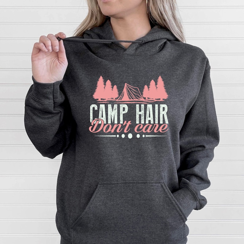 K&ouml;nnte beinhalten: Dunkelgrauer Kapuzenpullover mit Fronttasche und Kordelzug. Die Grafik auf der Vorderseite zeigt ein rosa Zelt und B&auml;ume mit dem Text "CAMP HAIR Don't care."