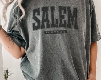 Puede incluir: Camiseta gris oscuro con la palabra "SALEM" en letras negras grandes y audaces en el pecho. Debajo de la palabra "SALEM" está la palabra "MASSACHUSETTS" en letras negras más pequeñas. La camiseta tiene un aspecto vintage y desgastado.