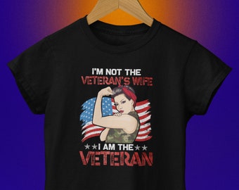 May include: Black t-shirt with the text "I'M NOT THE VETERAN'S WIFE I AM THE VETERAN" and an illustration of a woman flexing her bicep in front of an American flag design. The woman is wearing a red bandana and a camouflage top.